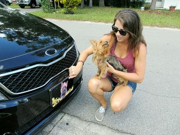 Young woman carrying yorkshire terrier while crouching by car on road