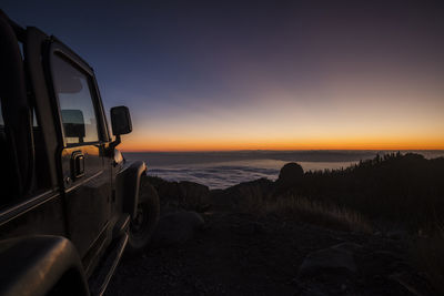 Vehicle parked by sea against sky during sunset