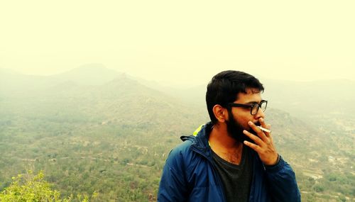 Man smoking cigarette while standing against mountain during foggy weather