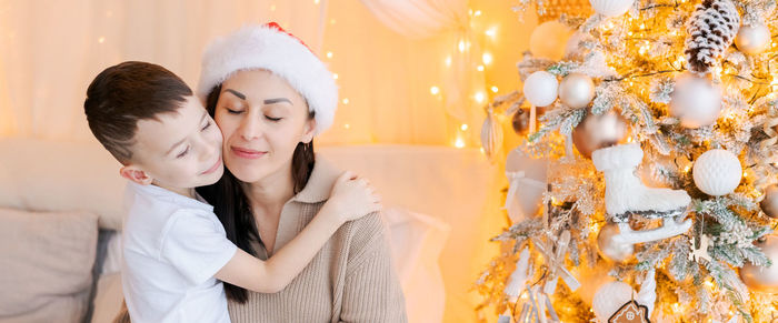Happy mom and son wearing santa claus hat cuddle on bed in decorated bedroom