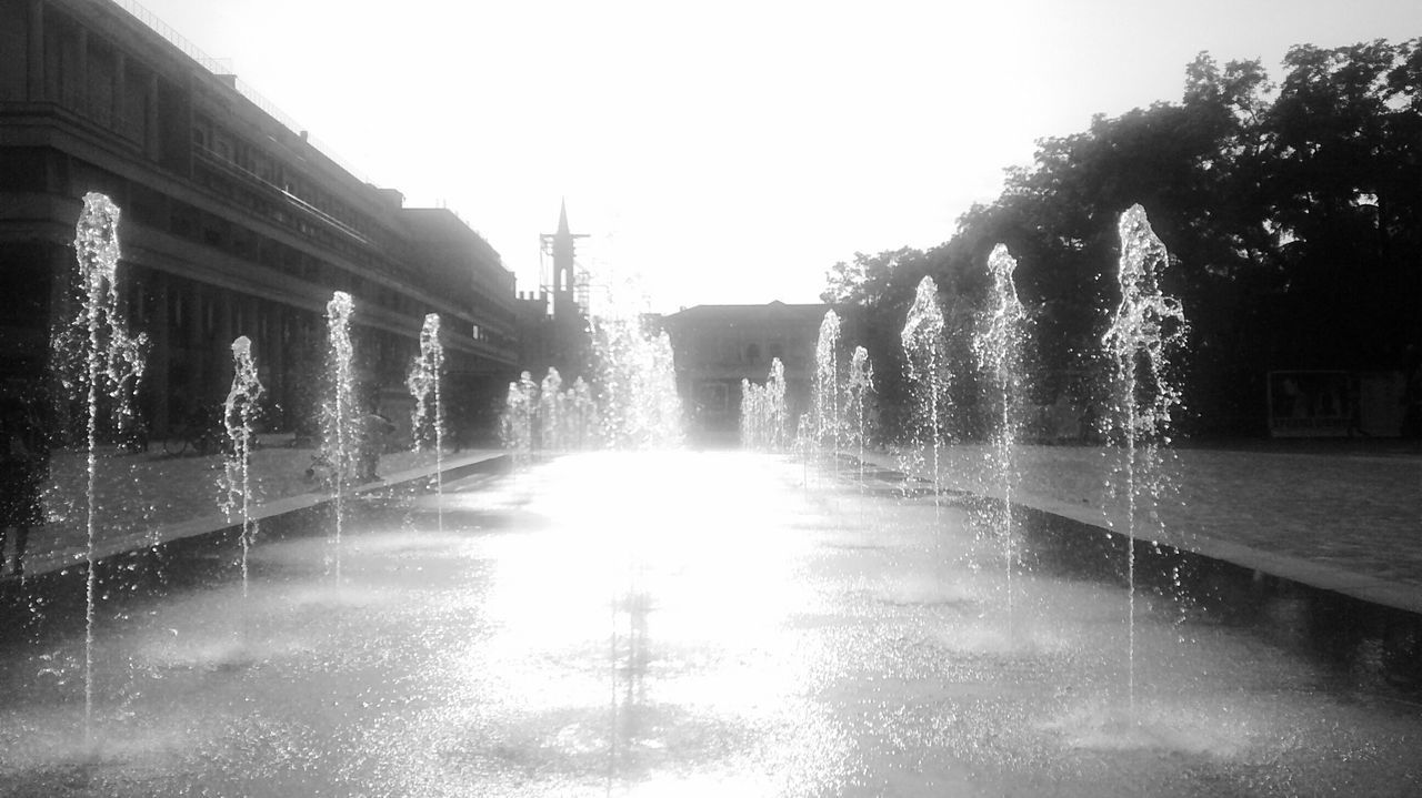 water, built structure, architecture, building exterior, clear sky, fountain, wet, reflection, sunlight, tree, motion, building, street, city, outdoors, street light, sunbeam, waterfront, rain, day