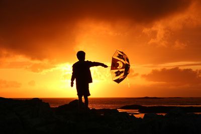 Silhouette boy holding umbrella while standing on rock against orange cloudy sky