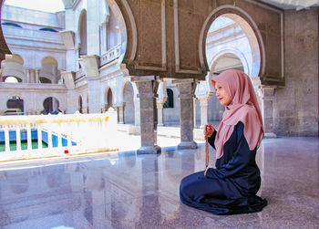 Full length of woman in hijab holding counting rosary beads while praying at mosque