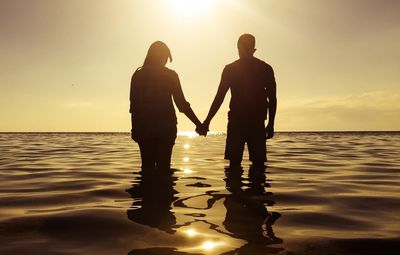 Silhouette couple holding hands while standing in sea against sky during sunset