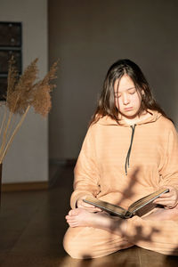 A girl in a beige tracksuit sits on the floor in a lotus position and reading a book