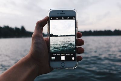 Cropped hand of person photographing lake with mobile phone against sky