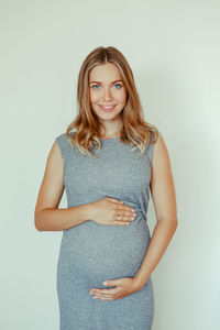 Portrait of pregnant young woman touching abdomen while standing against white background