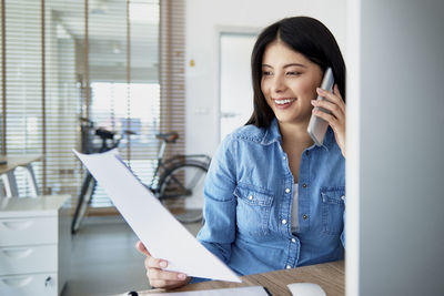 Smiling young woman holding document talking over mobile phone in office