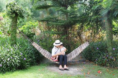 Full length of mother sitting with son on hammock amidst plants at park