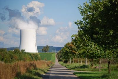 Cooling tower emitting smoke by country road against sky