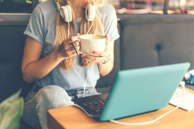  woman with headphones and a laptop at a table in a coffee shop holding a cup with a drink