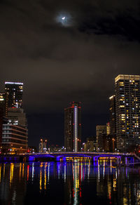 Illuminated buildings by river against sky at night