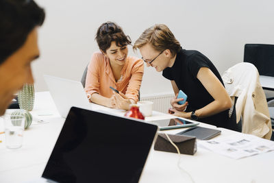 Smiling businesswoman sharing smart phone with transgender businessman while sitting in board room during meeting