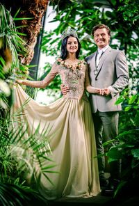 Portrait of smiling bride and groom holding hands while standing amidst plants