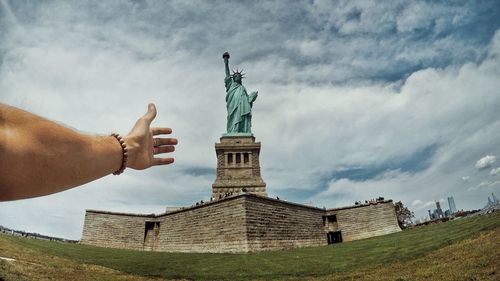 Low angle view of hand gesturing with statue of liberty in background against cloudy sky