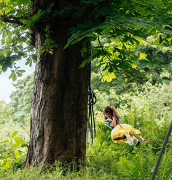 A young girl in a princess outfit smiles as she swings from a tree in the forest