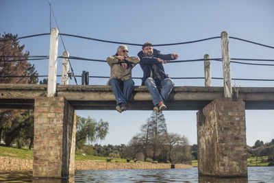 Senior man and adult son sitting on a bridge fishing together