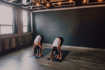 Fit barefoot unrecognizable women in sportswear concentrating and doing downward facing dog exercise on sports mats on wooden floor against white walls of spacious hall