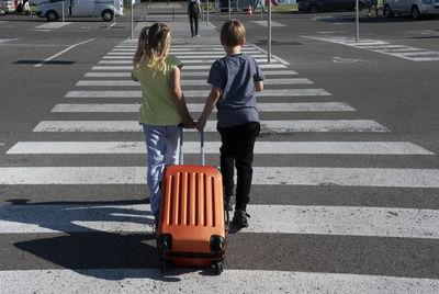 Rear view of siblings pulling suitcase on zebra crossing in city