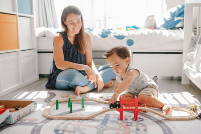 Mother and toddler boy playing with car wooden railway on floor at home. early age education 