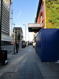 Man walking on footpath by street against sky in city