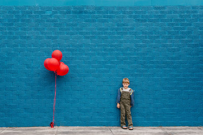 Full length of boy with red balloons standing against blue wall