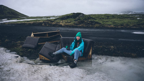 Portrait of woman sitting on abandoned sofa against sky during winter