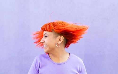 Excited adult female with closed eyes shaking bright ginger dyed hair against violet wall on street