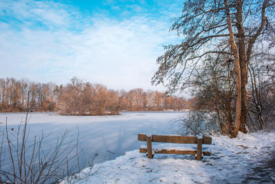 Bench by frozen lake against sky during winter