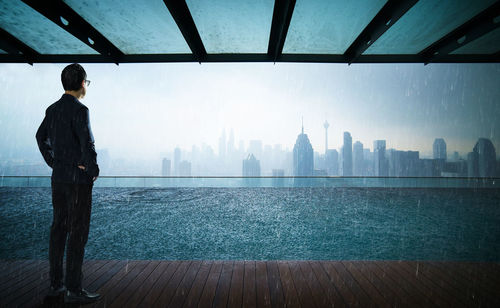 Rear view of businessman standing on pier by river against cityscape during monsoon