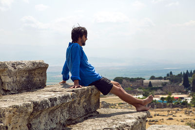 Man in a blue linen shirt and shorts sits on an old stone staircase colosseum in pamukkale turkey