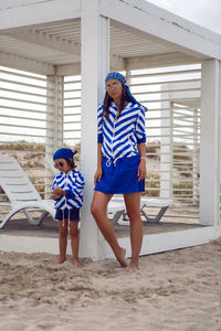Mother and her baby son in striped blue jackets walk along the beach next to a wooden gazebo