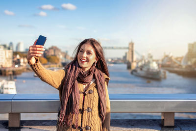 Smiling young woman taking selfie while standing by railing in city