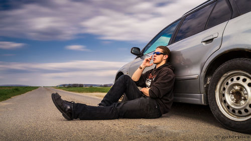 Full length of man smoking cigarette while relaxing by car on road
