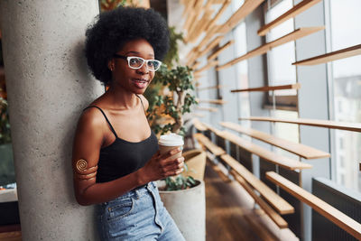 African girl with curly black hair and in casual clothes standing indoors with cup of drink.