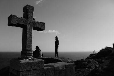 People on rock by religious cross against sky