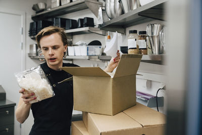 Young barista looking at package while standing by cardboard box in kitchen at cafeteria