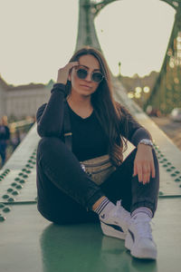 Portrait of young woman wearing sunglasses while sitting on the bridge