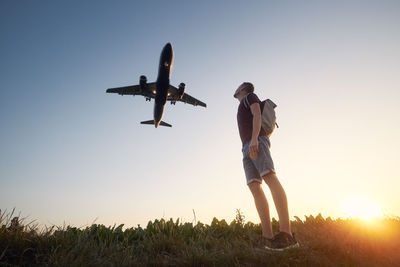 Man with backpack looking up to airplane landing at airport during beautiful summer sunset.