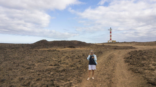 Full length of senior woman standing on field against lighthouse and sky