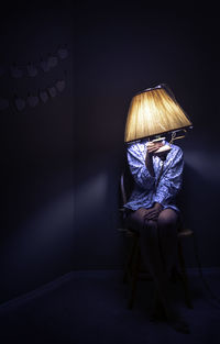 Young woman holding illuminated electric lamp while sitting on chair in darkroom at home