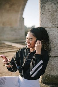 Happy female athlete adjusting in-ear headphones while using mobile phone on sunny day