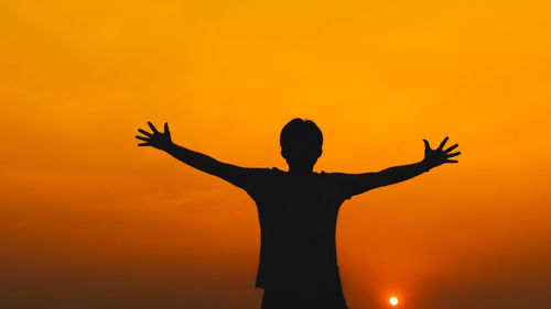 Silhouette man with arms outstretched standing against orange sky