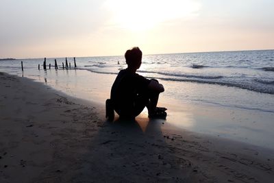 Man sitting at beach against sky during sunset