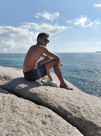 Shirtless man sitting by sea on rock during sunny day