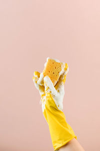 Person in rubber glove holds sponge with foam washing dishes on beige closeup