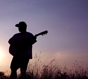 Silhouette man playing guitar on field against sky at sunset