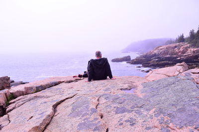 Rear view of man sitting on rock by sea against clear sky