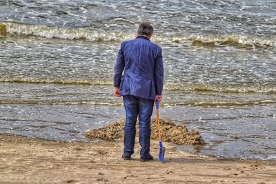 Rear view of man holding shovel while standing on sand against sea at beach