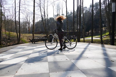 Woman with bicycle tossing hair on checked footpath at park